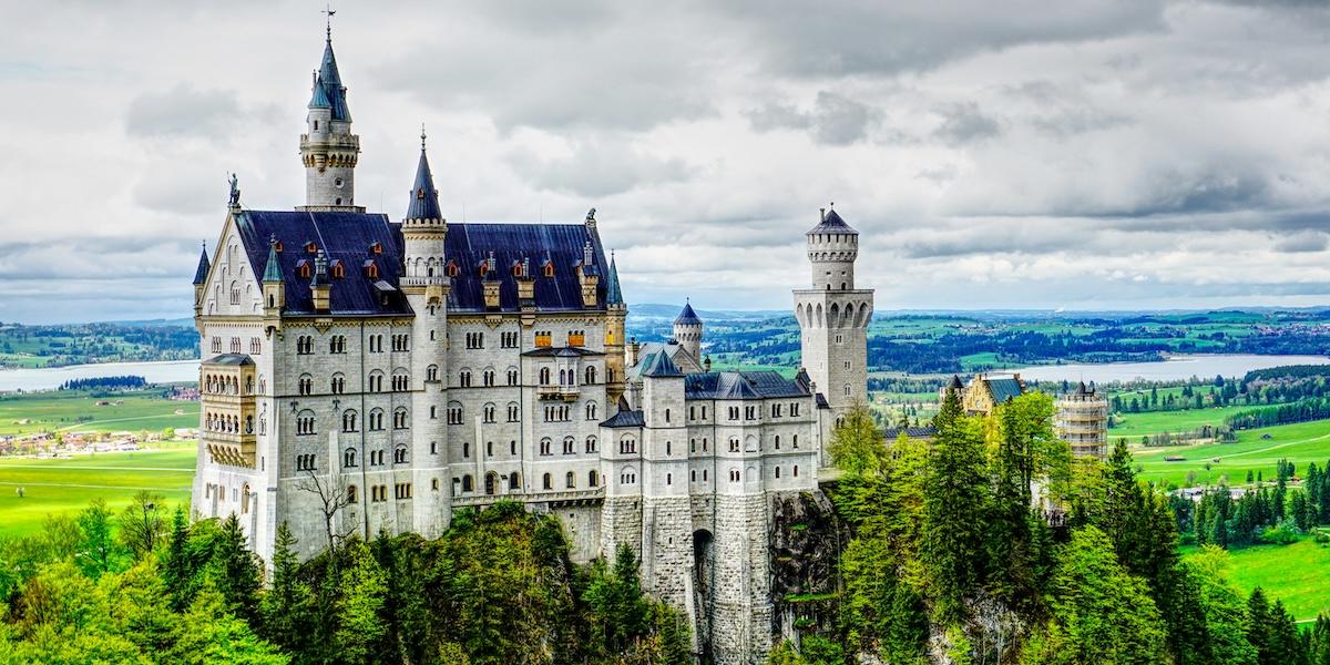 Luftig vy över Neuschwanstein Castle i de bayerska Alperna med torn, spiror och skogslandskap i bakgrunden.