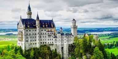 Luftig vy över Neuschwanstein Castle i de bayerska Alperna med torn, spiror och skogslandskap i bakgrunden.