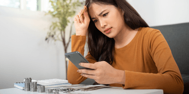 Ung person granskar ekonomi i mobilen med orolig min, omgiven av mynt och budgetpapper.