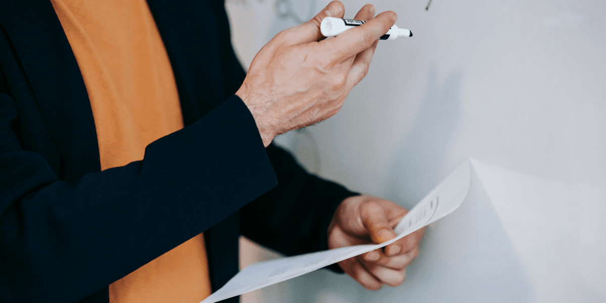 Man skriver på whiteboard med papper i handen – symboliserar lärare i klassrum.