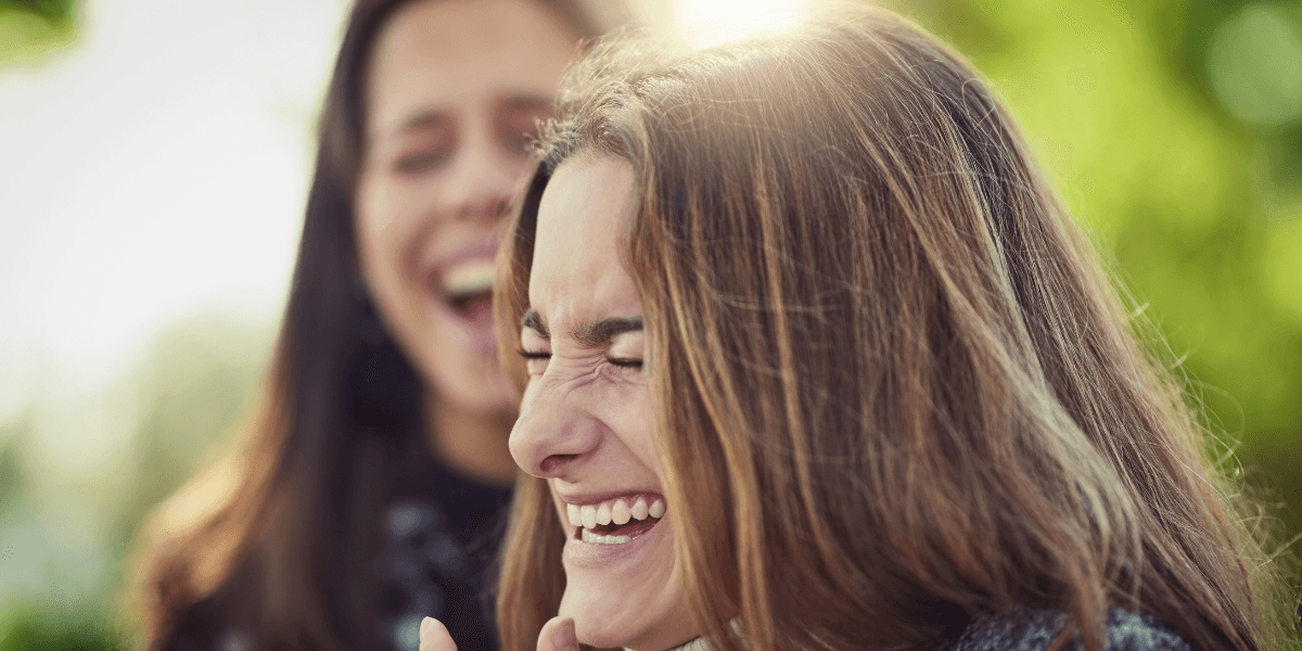 Två kvinnor skrattar tillsammans utomhus i ett grönt, soligt miljö – en symbol för laughter yoga och glädjefylld träning.