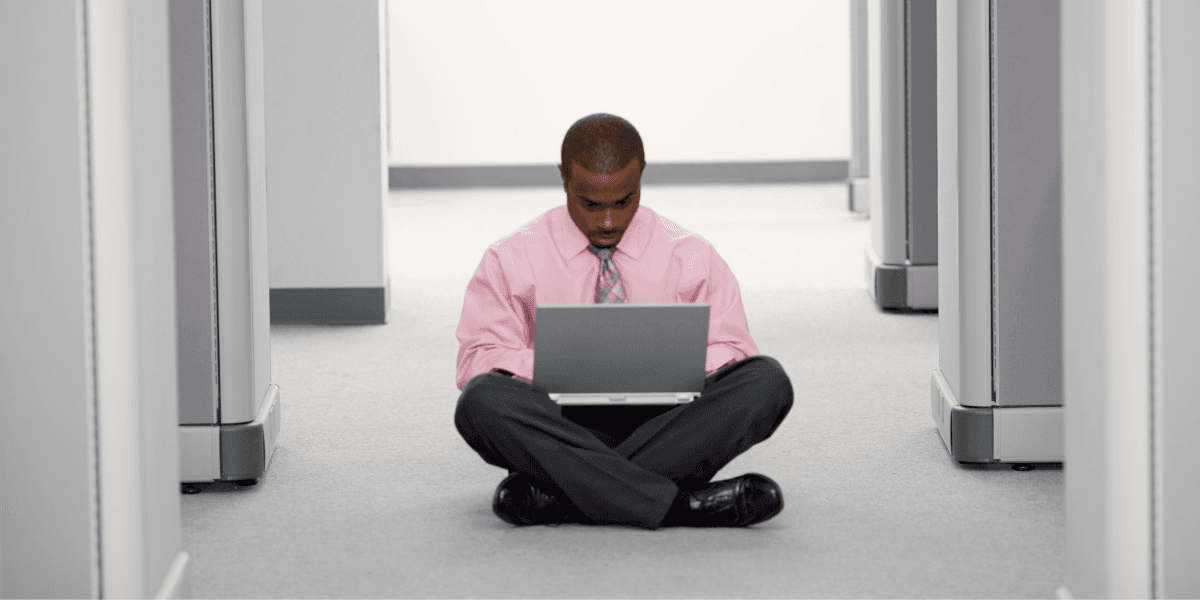 Anställd utan skrivbord sitter på golvet med dator – illustrerar platsbrist i kontorsmiljö.