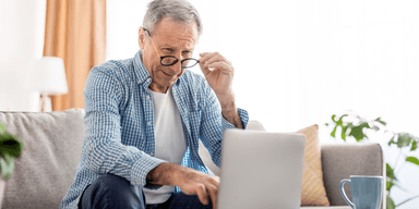 Äldre man sitter i vardagsrum och tittar på laptop, symboliserar pensionär som kollar sin pensionsutbetalning online.