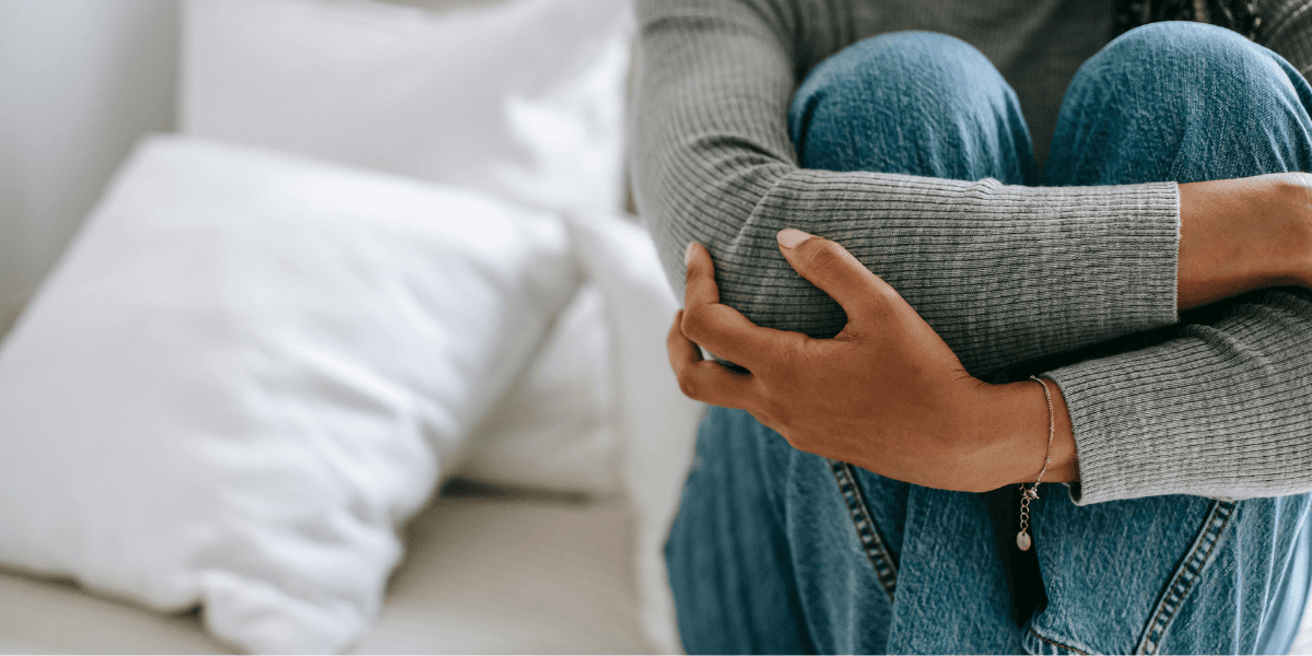 Ung person i grå tröja sitter hopkrupen på säng, symboliserar känslan av ensamhet bland unga vuxna.