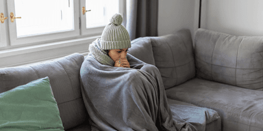 Person med mössa och filt sitter frusen på en soffa i ett kallt vardagsrum.